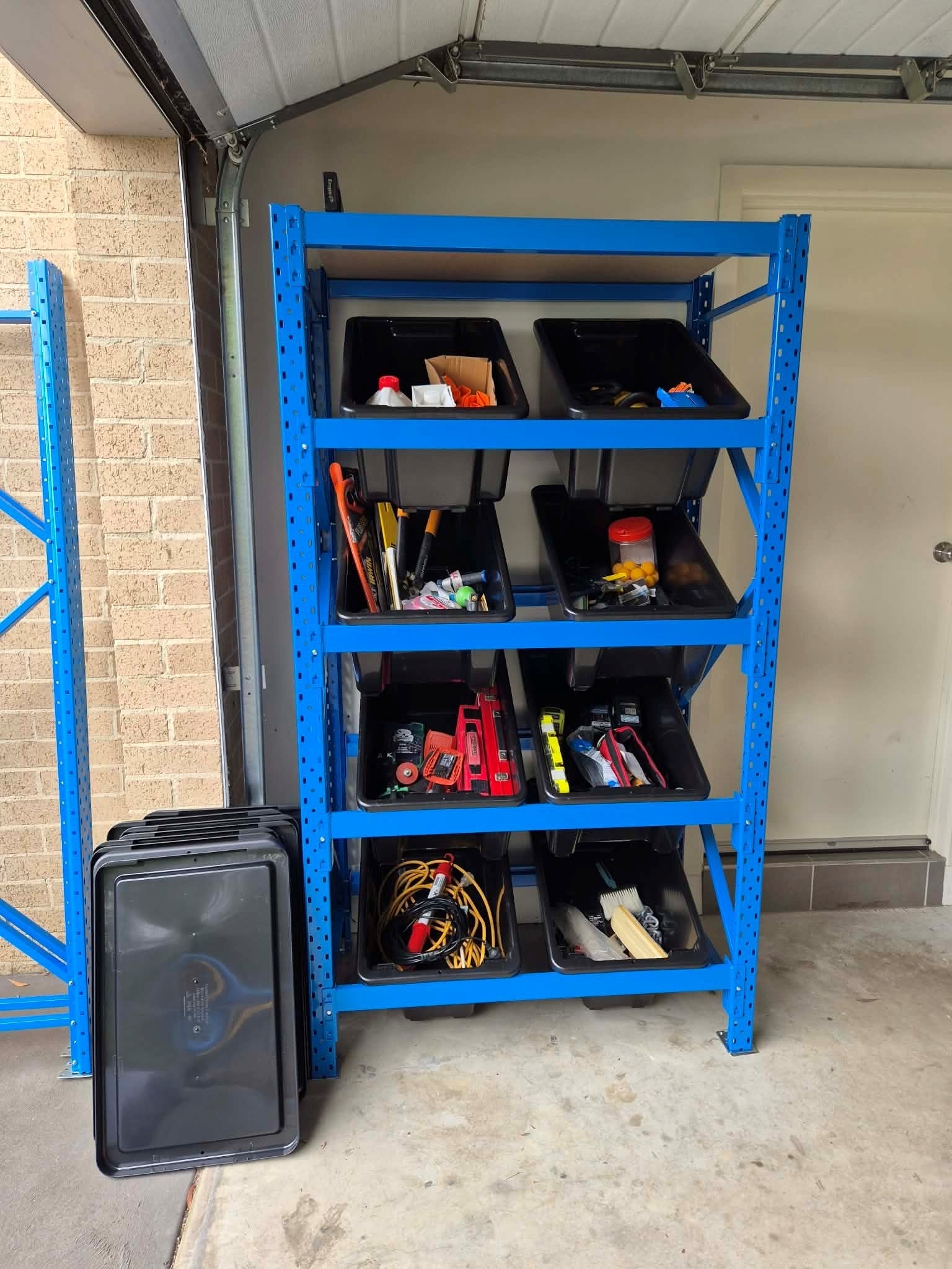 Blue steel garage shelving unit fitted with angled black storage bins, neatly organising hand tools, cables, and small hardware inside an open garage space.